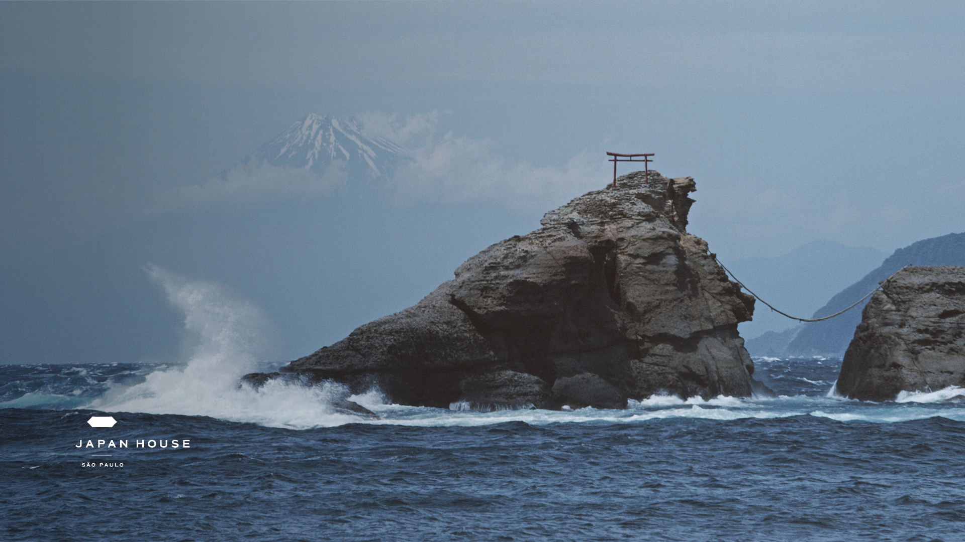 Onda batendo em rocha no meio do mar azul escuro. Ao fundo é possível enxergar a silhueta do Monte Fuji, no Japão, em meio às nuvens.