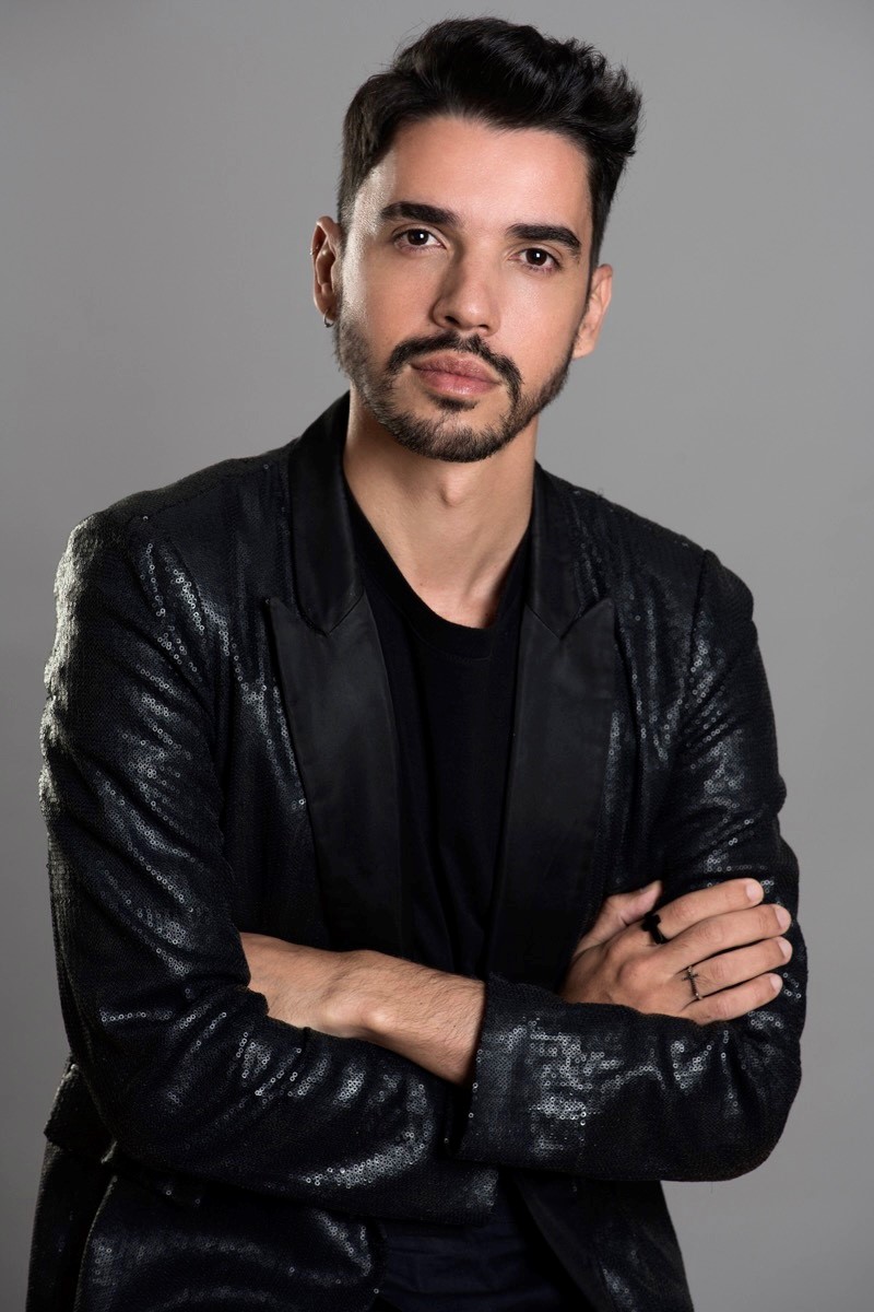 Tiago é um homem jovem, tem barba, cavanhaque e cabelos escuros. Ele usa uma camisa preta e uma jaqueta de couro preta. Tiago está com os braços cruzados e olha sério para frente. 
